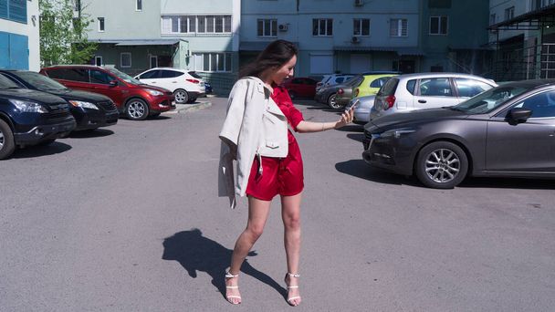 Emotional young beautiful woman walking on street on white high heels shoes and red dress.Beautiful brunette woman in shock with open mouth looking on phone. - Photo, image