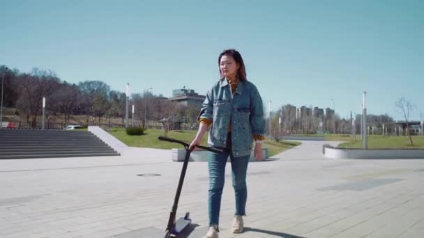 Nuori aasialainen nainen kävelee potku skootteri kesäpuistossa
 - Materiaali, video