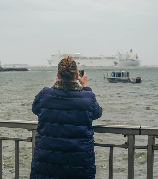 NEW YORK - APRIL 30, 2020: Spectators on the Brooklyn shoreline take pictures as the USNS Comfort Hospital Ship departing New York City after a month of caring for patients with the Coronavirus - Фото, изображение