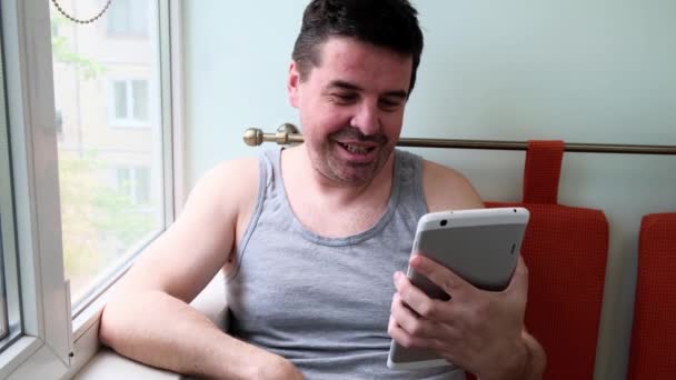 40 yaşında orta yaşlı bir adam evinin penceresinin yanında oturuyor, tablet bilgisayarla konuşuyor, kablosuz internet bağlantısı kullanıyor. İletişimciler karantinada ve kendilerini izole ediyorlar.. - Video, Çekim