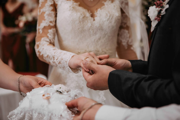 Ženich dává prsten na ruku své nevěstě během svatebního obřadu na matrice. Nevěsta v bílých elegantních krajkových šatech a ženich v černém obleku. Rodiče slouží prsten - Fotografie, Obrázek