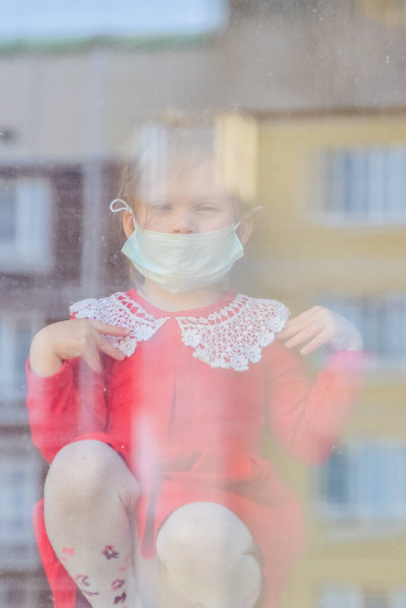 the child sits at the window in a medical mask and looks out the window - Φωτογραφία, εικόνα