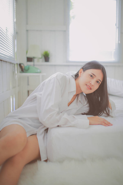 Gyönyörű fiatal ázsiai nő ül a fehér ágynemű lágy reggeli fényben. - Fotó, kép