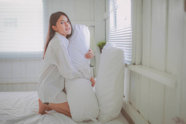Портрет жінки - азіата, яка стоїть на колінах на ліжку після пробудження вранці в спальні з сонячним світлом, що світить крізь штори..   - Фото, зображення