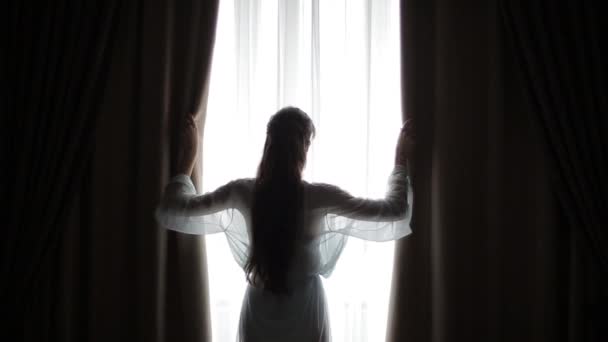 A fiatal nő felemeli a kezét és áthajt a függönyön a hálószobában, megvédve magát a reggeli fénytől, ami az ablakon keresztül jön be. Egy gyönyörű lány sziluettje elhúzza a függönyt az ablakon. - Felvétel, videó
