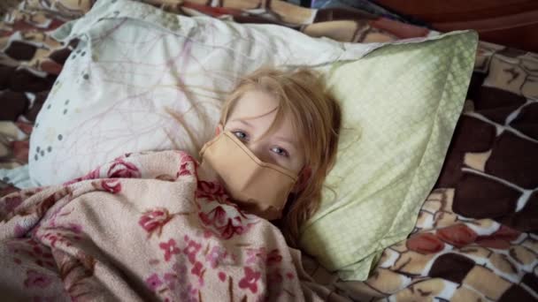Little girl is sick. Lying on a bed in a medical mask, covered with a blanket. - Footage, Video