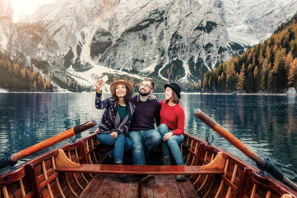 Happy young cheerful friends students have an adventure in nature and travel in a wooden boat on a mountain lake. Travel blogger taking selfie photos for social network - Photo, Image