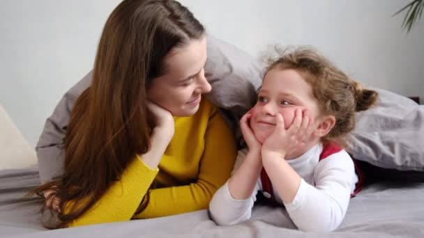 Portrait of cute little child kid daughter and cheerful young mother lying on bed covered with blanket look at camera posing at home, happy family mom and small girl spend time together - Imágenes, Vídeo