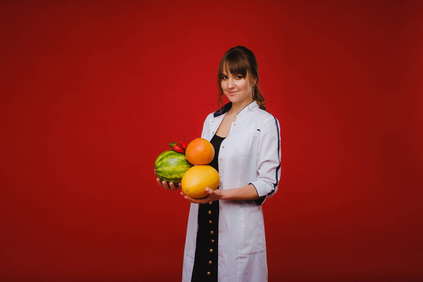 a female doctor nurse in a white coat with fruit in her hands poses on a red background, melon, watermelon, strawberry and grapefruit. - Zdjęcie, obraz