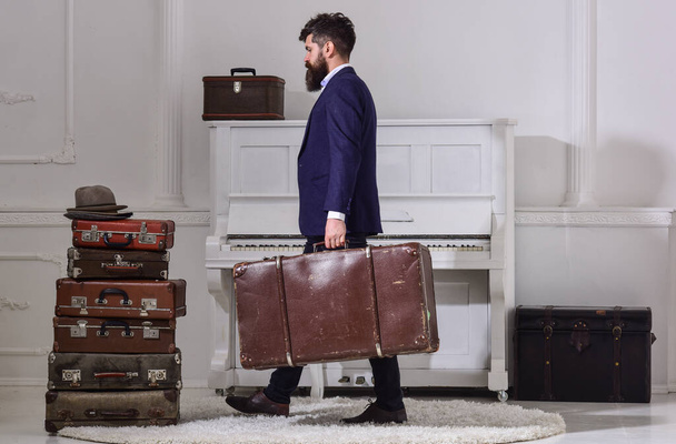 Macho stylish on strict face walks and carries big vintage suitcase, side view. Man, traveller with beard and mustache with baggage, luxury white interior background. Baggage and travelling concept - Fotografie, Obrázek