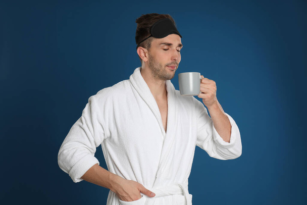 Young man in bathrobe with cup of coffee on blue background - Foto, afbeelding