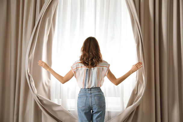 Woman opening window curtains at home in morning, back view - Foto, Bild