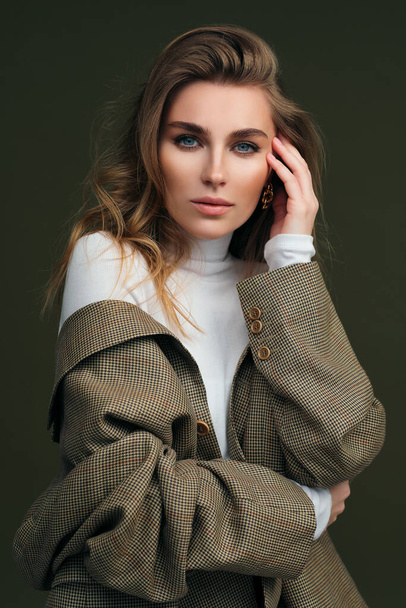 A young beautiful blonde woman in a white turtleneck and a mans jacket poses on a green olive background in the studio. - Foto, afbeelding