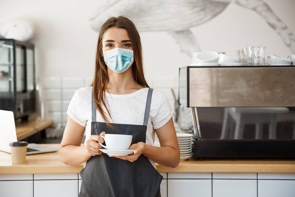 Business Owner Concept - Beautiful Caucasian Barista in face mask offers hot coffee at the modern coffee shop - Photo, Image