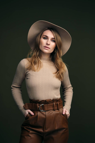 A young attractive blonde woman in a beige sweater, fedora hat and brown leather trousers poses on a dark green olive background. Studio portrait of a beautiful girl. - Fotó, kép