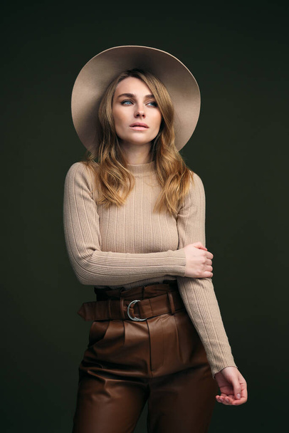 A young attractive blonde woman in a beige sweater, fedora hat and brown leather trousers poses on a dark green olive background. Studio portrait of a beautiful girl. - 写真・画像