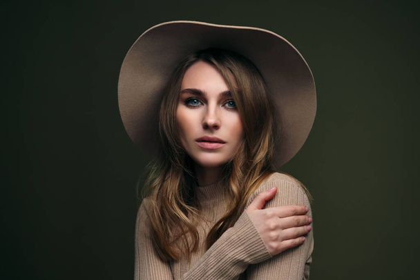 A young attractive blonde woman in a beige sweater, fedora hat and brown leather trousers poses on a dark green olive background. Studio portrait of a beautiful girl. - Foto, Imagem