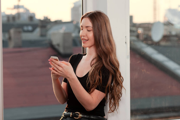Smiling caucasian woman is sitting on the windowsill and writing message on smartphone at home. Side view of young woman using smartphone to connect to internet and use social media with happy. - Fotoğraf, Görsel