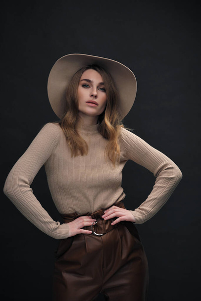 A young sensual sexy woman with a beautiful face and clear natural skin poses in the studio against a black background. Beige slinky sweater and hat with fields. - Фото, изображение