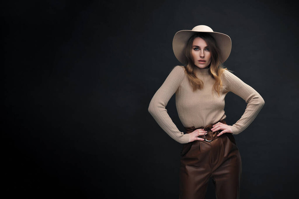 A young sensual sexy woman with a beautiful face and clear natural skin poses in the studio against a black background. Beige slinky sweater and hat with fields. - Foto, immagini