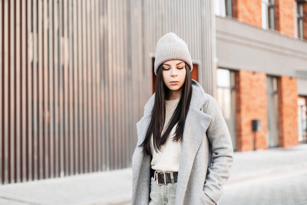 ヴィンテージコートのスタイリッシュなニット帽のかなりファッション若いブルネットの女性が立って、明るい春の日に近代的な建物の近くの道路を見下ろしています。流行の服のモデル都市の女の子。カジュアルルック. - 写真・画像