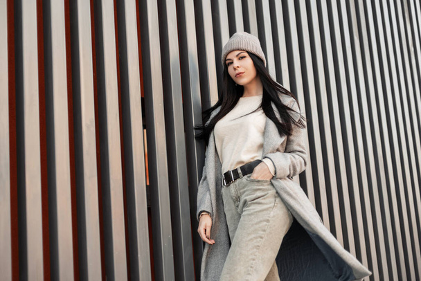 Young fashionable brunette woman with long hair in stylish gray coat in jeans in knitted trendy hat posing near vintage metal building. Urban attractive girl fashion model. Street casual style. Spring - Photo, Image