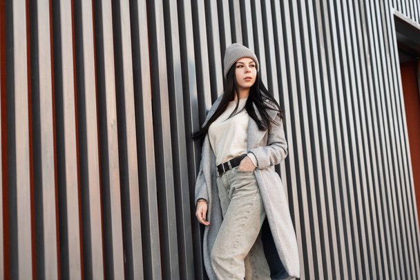 Stylish pretty young woman with long black hair in trendy knitted hat in vintage spring coat in jeans in sweater poses outdoors on bright day. Attractive brunette girl fashion model near metal wall. - 写真・画像