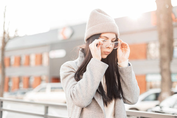 Young stylish beautiful hipster woman in fashionable knitted hat in vintage coat puts on trendy glasses in the city on the background bright spring sunset. Attractive urban girl model posing outdoors. - Фото, изображение