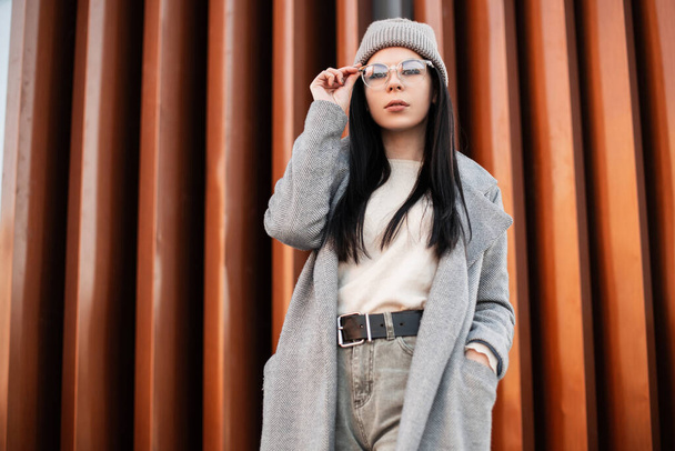 Melko tyylikäs nuori hipster nainen muodikas takki neulottu harmaa hattu vintage lasit poseeraa lähellä metalliseinää kaupungissa. Kaupunkien Euroopan trendikäs tyttö malli kadulla. Kevään rento tyyli
 - Valokuva, kuva