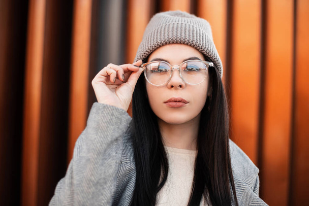 Portré gyönyörű fiatal nő hipster hosszú haj szexi ajkak elegáns kötött kalap divatos kabát közelében modern fal szabadban. Elég divatos lány egyenesíti divat szemüveg az utcán. - Fotó, kép