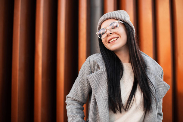 Πορτρέτο χαρούμενη νεαρή γυναίκα με όμορφο χαμόγελο σε κομψά γυαλιά σε μοντέρνο πλεκτό καπέλο σε vintage παλτό στο δρόμο κοντά σε μεταλλικό τοίχο. Χαριτωμένο μοντέρνο ευτυχισμένη hipster κορίτσι θέτει και χαμογελώντας σε εξωτερικούς χώρους - Φωτογραφία, εικόνα