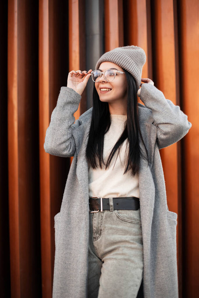 Beautiful cheerful young hipster woman with cute smile in fashionable clothes straightens glasses. Fashion portrait happy girl model in sweater in knitted fashionable hat in trendy coat in the street. - Foto, Bild