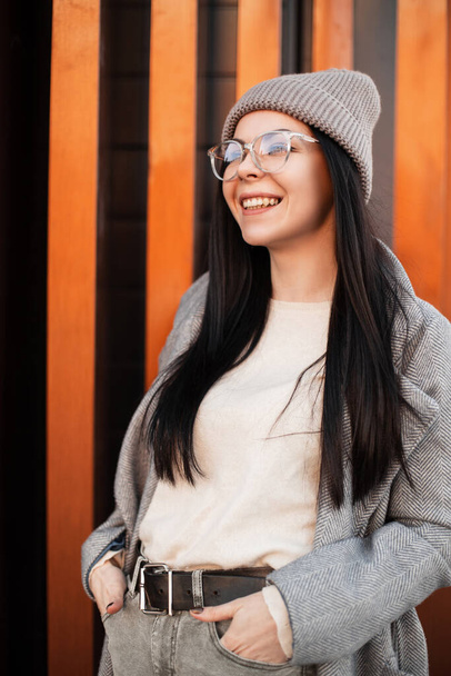 Happy attractive young woman with smile in fashionable knitted hat in stylish coat in fashion glasses poses outdoors in the city near a metal wall. Joyful trendy hipster girl is smiling on the street. - Photo, Image