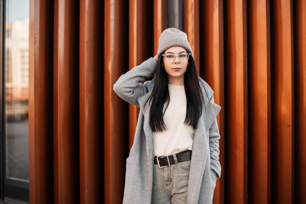 Attractive young woman in fashionable coat in stylish knitted hat in trendy glasses in sweater poses near vintage metal wall in the city. Hipster urban girl outdoors. Spring fashion. New collection. - Φωτογραφία, εικόνα