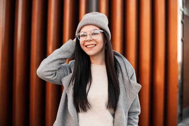 Positive young hipster woman in fashion coat in fashionable glasses straightens knitted trendy hat and funny smiles near metal wall. Pretty stylish girl with charming smile on the street in the city - Photo, Image