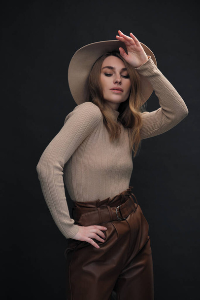 A young sensual sexy woman with a beautiful face and clear natural skin poses in the studio against a black background. Beige slinky sweater and hat with fields. - Valokuva, kuva