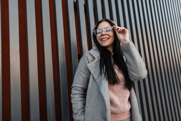 Gelukkige jonge vrouw hipster rechtzet trendy bril en positieve glimlach. Fris portret schattig vrolijk meisje met mooie glimlach in grijs modieuze jas in vintage roze hoodie in de buurt van metalen muur in de stad. - Foto, afbeelding