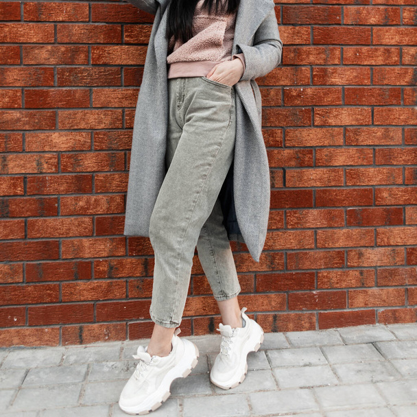 Moderne jonge vrouw in stijlvolle roze sweatshirt in vintage jeans in grijze lange jas in modieuze sneakers staat in de buurt van bakstenen muur. Stedelijk meisje in casual trendy kleren. Straatstijl. Voorjaar look.Close-up - Foto, afbeelding