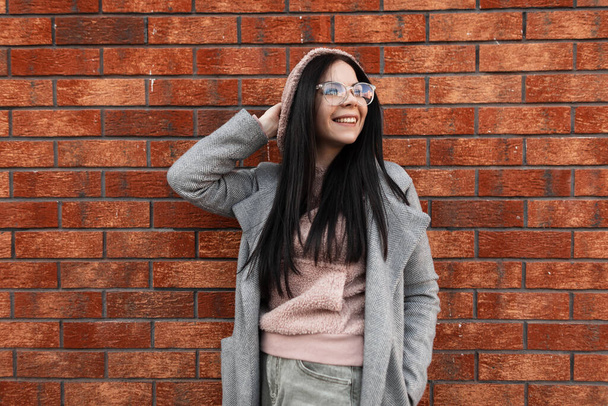 Onnellinen söpö nuori hipster nainen positiivinen hymy trendikäs vaaleanpunainen huppari tyylikäs harmaa takki muodikas lasit poseeraa ja hymyilee lähellä tiili vintage seinä kaupungissa. Iloinen tyttö kadulla
. - Valokuva, kuva