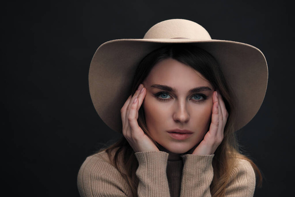 A young sensual sexy woman with a beautiful face and clear natural skin poses in the studio against a black background. Beige slinky sweater and hat with fields. - Foto, imagen