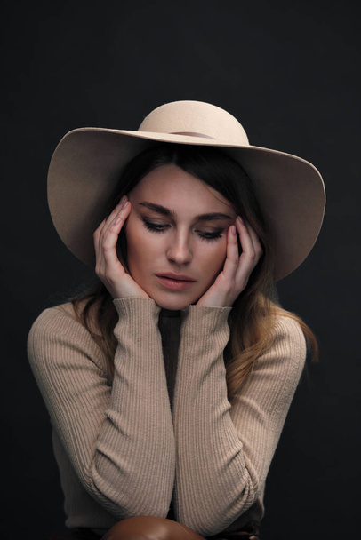 A young sensual sexy woman with a beautiful face and clear natural skin poses in the studio against a black background. Beige slinky sweater and hat with fields. - Фото, зображення
