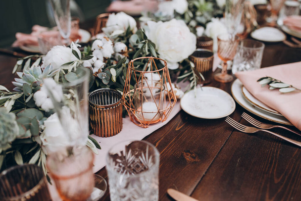 Wedding decor. On wooden banquet table are glasses, plates, candles, table is decorated with compositions of cotton and eucalyptus branches, plates are decorated with napkins and sprig of Italian greenery - 写真・画像