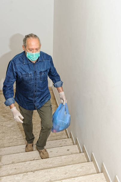 Wegen der Coronavirus-Infektion COVID-19 liefern wir einige Lebensmittel zur Quarantäne nach Hause. Mann mit Schutzhandschuhen steigt Treppe hinauf und hält Einkaufstasche in der Hand - Foto, Bild