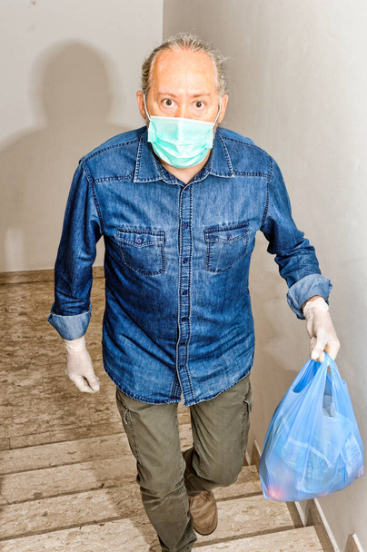 σπίτι παραδίδοντας κάποια είδη παντοπωλείου στο χρόνο καραντίνας λόγω της μόλυνσης από τον ιό της στέψης COVID-19. Άντρας με προστατευτικά γάντια, ανεβαίνει σκάλες κρατώντας μια τσάντα παντοπωλείου - Φωτογραφία, εικόνα