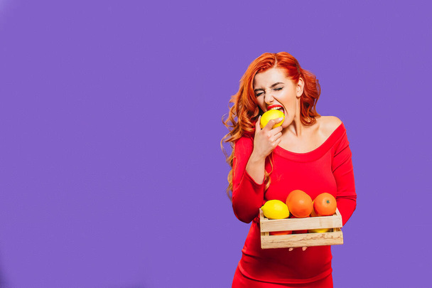Jong mooi meisje heeft een mand met sinaasappels en citroenen. Een meisje met rood haar in een rode jurk bijt een citroen op een paarse achtergrond. Een spandoek. Een plek voor sms 'jes. Voedingsmiddelen van het coronavirus. - Foto, afbeelding
