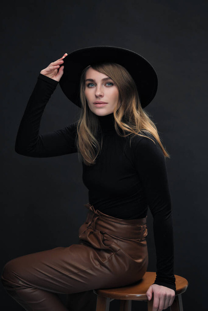 Portrait of young beautiful blond woman with light makeup and blue eyes touching her face. Dark background. Black tight sweater and hat with fields. - Photo, Image