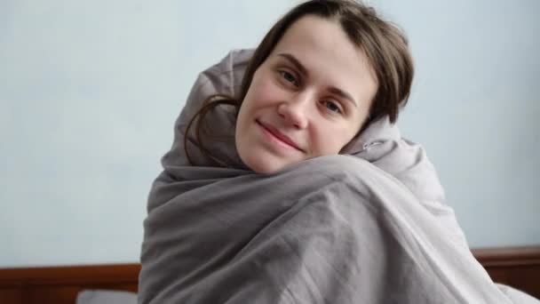 Portrait of smiling cheerful young woman relaxing in comfortable cozy bed looking at camera, cute girl having fun posing covering grey soft warm blanket - Felvétel, videó