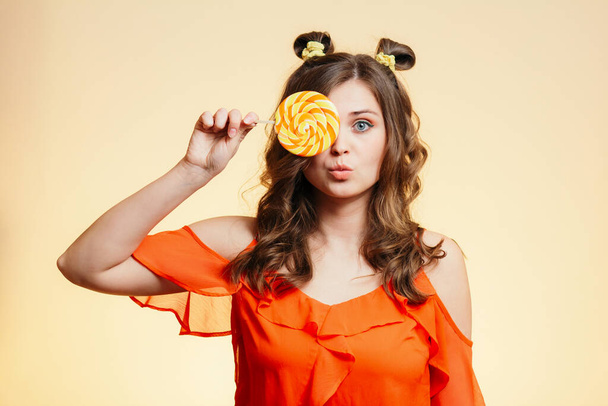 portrait of cute funny girl grimacing with a round caramel on orange background, young woman with a striped candy, concept of sweets, positive mood - Fotó, kép
