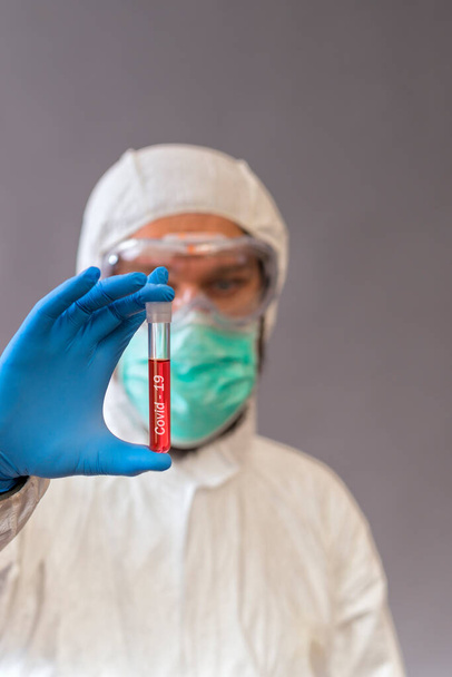 Male doctor with surgical mask, goggles and protective suit holding blood samples on gray backround. Vials with cure Covid-19. - Foto, afbeelding