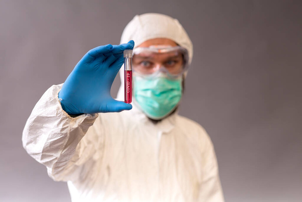 Male doctor with surgical mask, goggles and protective suit holding blood samples on gray backround. Vials with cure Covid-19. - Photo, Image